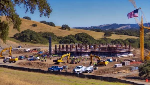 An expansive view of a Novato, California construction project surrounded by golden hills. Workers and machinery, including excavators and dump trucks, are actively building a modern structure in harmony with the serene landscape.