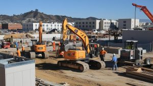 A construction site in Paramount, California, featuring multiple excavators and workers wearing safety gear. The background includes modern buildings and scenic hills, showcasing an active development project.