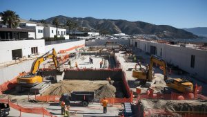 A large construction project in Paramount, California, with several excavators and construction workers building a foundation. The site is surrounded by safety fencing, modern structures, and mountainous terrain in the distance.