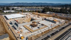 Aerial view of a construction site in Livermore, California, with foundational work in progress, cranes, and surrounding roads, set against a backdrop of mountains and greenery.