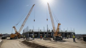 Active construction site in Martinez, California, featuring two large cranes, workers, and a partially built structure under a bright blue sky.