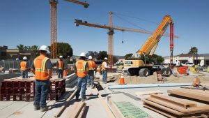 Construction workers in Monrovia, California, at a busy building site with cranes and heavy machinery under a bright blue sky.