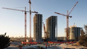 A construction site at sunrise or sunset with tall, partially constructed buildings flanked by several cranes. Workers in safety gear are spread out across the site, adding a sense of scale and progress to the construction of high-rise structures.