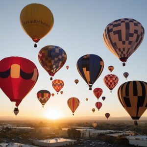 an image showcasing a colorful hot air balloon festival at sunrise in Vacaville, CA. Include multiple balloons of various shapes and sizes soaring above the city, with the sun peeking over the horizon.