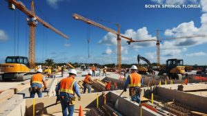 Construction workers on-site in Bonita Springs, Florida, working on a major development project with cranes and heavy equipment in the background under a bright blue sky.

