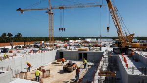 Active construction site in Fort Pierce, Florida, featuring multiple cranes, workers in safety gear, and partially built structures under a clear blue sky.