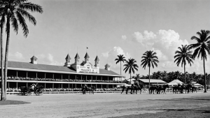 A vintage black-and-white photo of Hialeah Park in Florida, featuring palm trees, a grandstand with spectators, and horse-drawn carriages, evoking its historic charm.