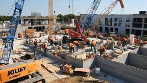 Active construction site in Joliet, Illinois with workers building structures, cranes in operation, and equipment labeled 'Joliet' in the foreground.