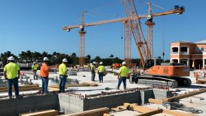 Construction workers in safety vests and helmets working on a project in Cape Coral, Florida, surrounded by cranes, building equipment, and a bright blue sky.