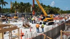 Construction workers and heavy equipment on a large job site surrounded by palm trees in Coconut Creek, Florida, showcasing an active infrastructure project.