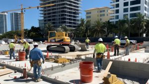 Construction workers at a building site in Coral Gables, Florida, featuring a yellow excavator and modern high-rise buildings in the background, illustrating active urban development.