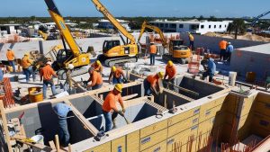 A group of construction workers assembling wooden structures on a sunny day in Deerfield Beach, Florida. The site includes cranes and partially constructed buildings, showcasing active project progress.