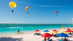 A lively beach scene in Pompano Beach, Florida, with parasailers, paddleboarders, and colorful umbrellas along a turquoise shoreline.