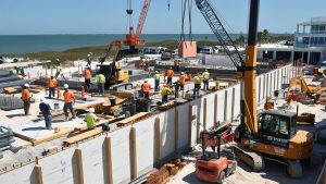 A bustling construction site near the ocean in Pompano Beach, Florida, featuring workers, heavy machinery, and cranes under a clear blue sky.