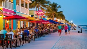 Colorful beachside restaurants with outdoor seating in Pompano Beach, Florida, showing diners enjoying the evening under bright umbrellas and palm trees.