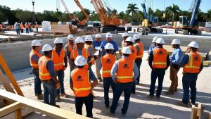 Construction crew in Pembroke Pines, Florida, gathered on-site in safety vests and helmets, reviewing blueprints with cranes and equipment in the background.