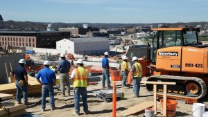 Construction workers wearing safety gear at a busy Waterbury, Connecticut construction site with heavy equipment and cityscape in the background.