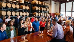 A lively bourbon tasting event in Louisville, Kentucky, showcasing a diverse group of people enjoying drinks at a distillery surrounded by stacked barrels.