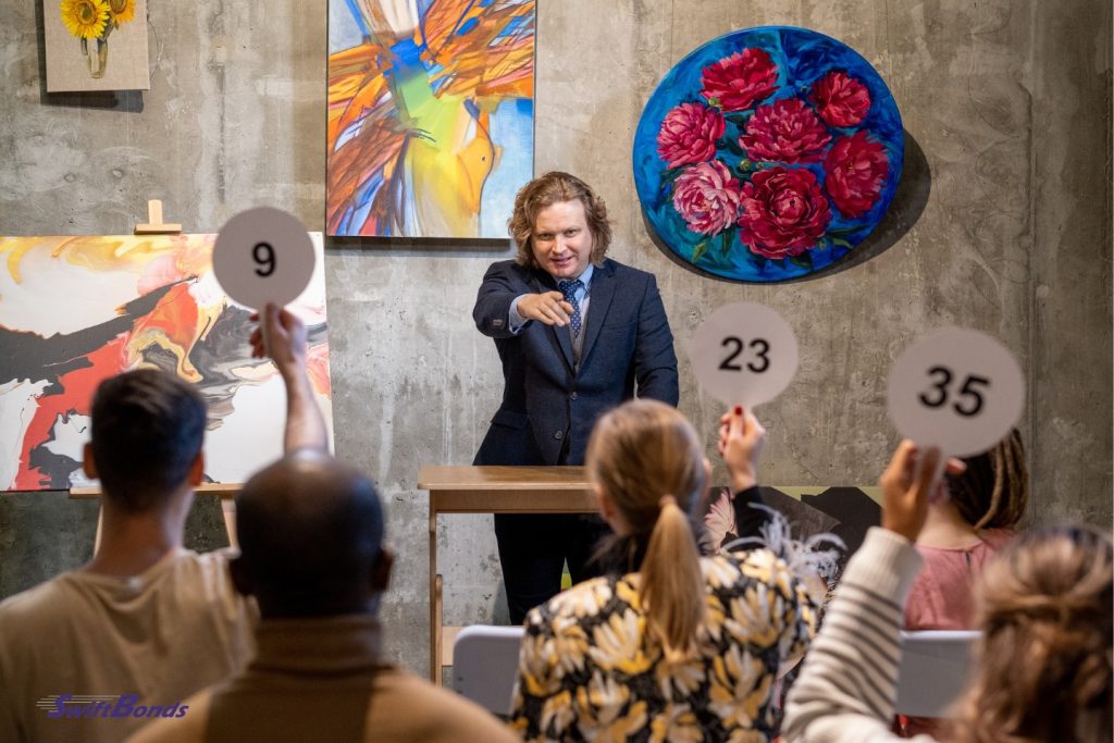 A male auctioneer gestures toward a bidder holding an auction paddle.