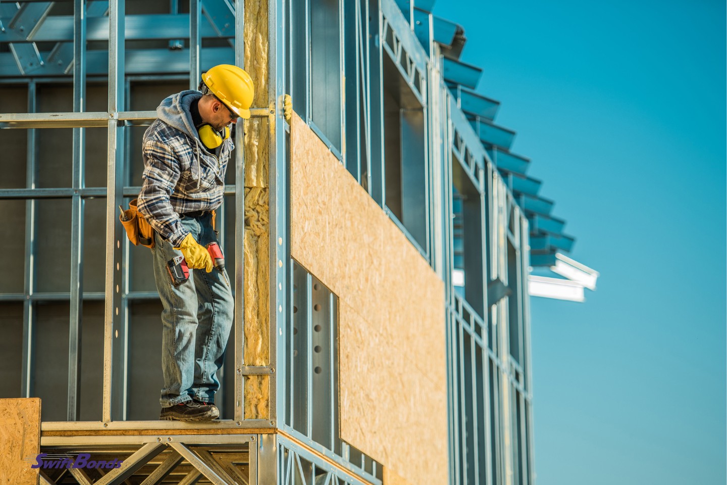 Standing atop the unfastened wall of the house's steel structure, a construction worker holds an electric cordless screwdriver.