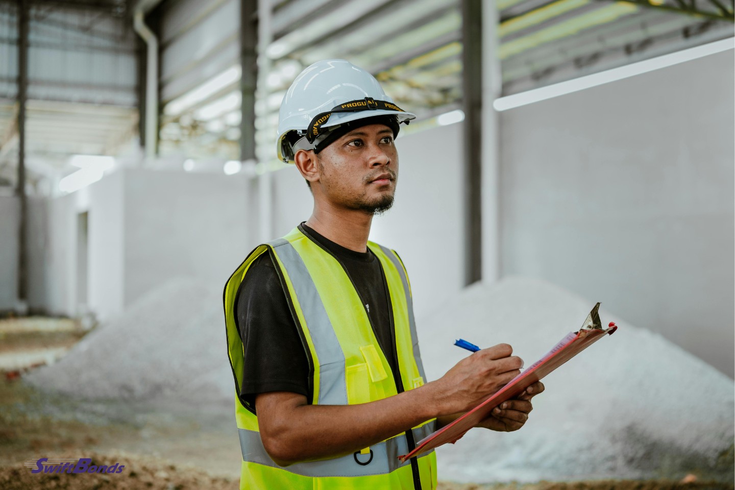 A construction worker with a pen and clipboard is shown in this portrait, with a white hard hat.