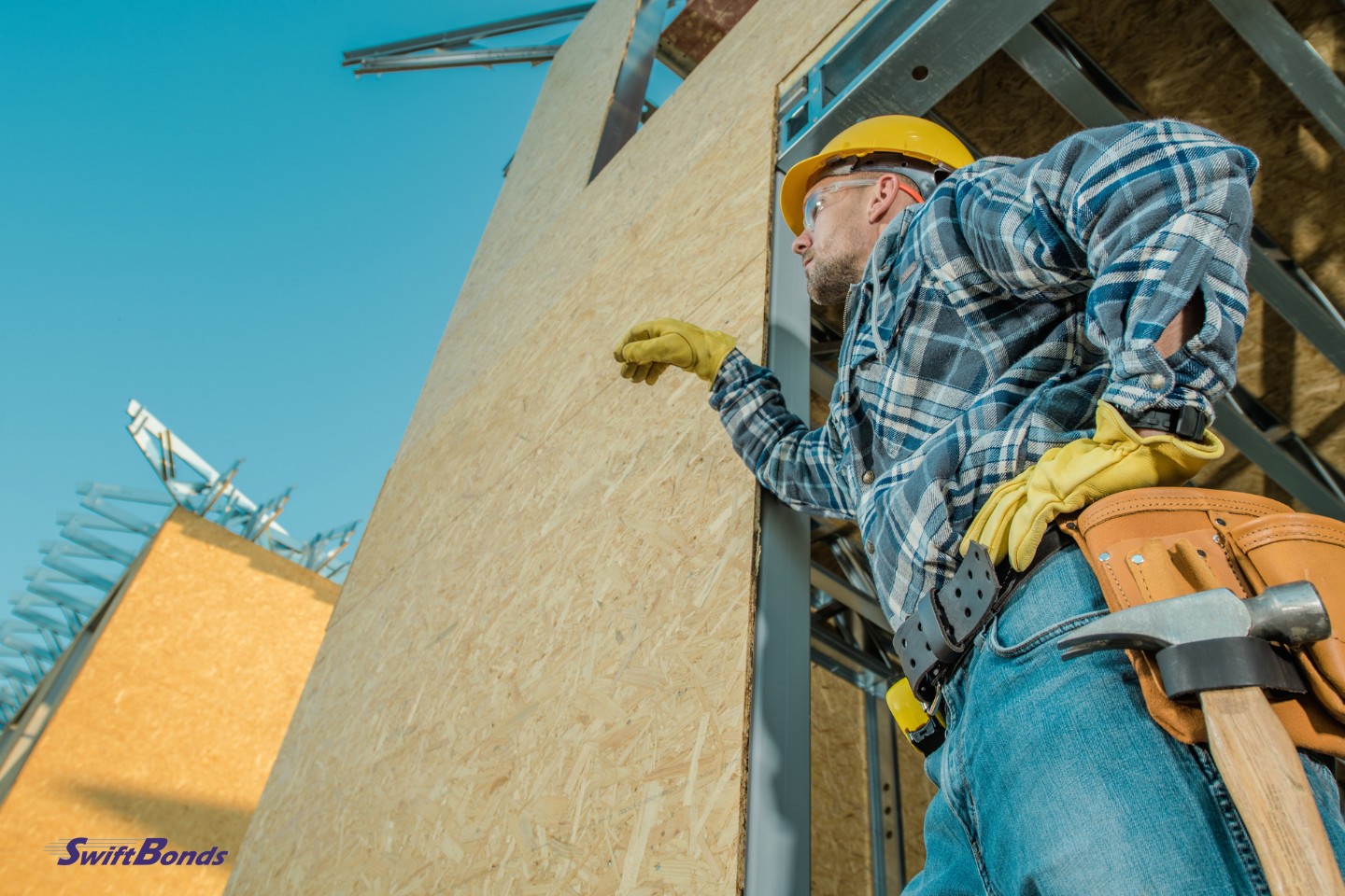 At the entryway of the built-up house stands a construction worker with his belt tools and yellow hard cap.