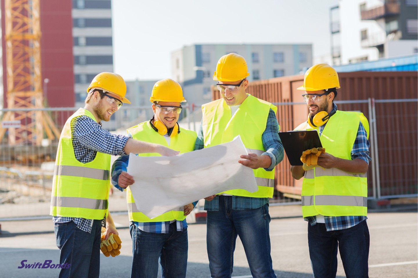 Outside, a group of happy builders wearing hard hats are holding a tablet PC and a blueprint.