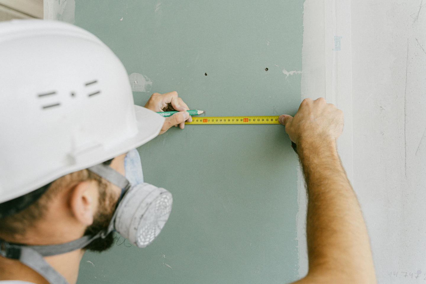 A worker wearing a hard hat and respirator mask measuring a wall with a tape measure.