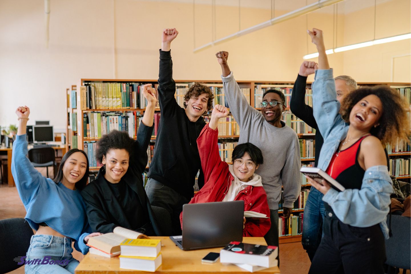 A group of students are standing next to a brown wooden table, raising their hands and grinning.