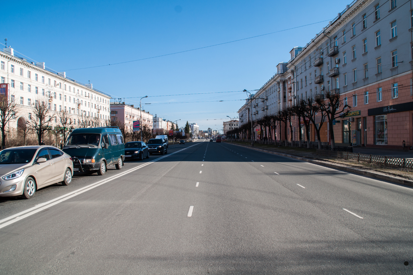 Wide city street with cars lined up along urban buildings under a clear sky.