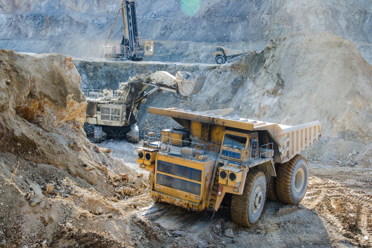 Heavy mining equipment, including an excavator and haul trucks, operating within a rocky open-pit mine.