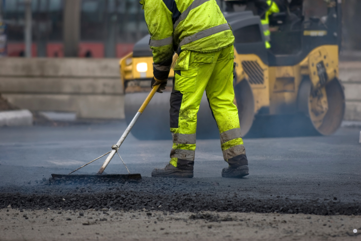 Road worker spreading fresh asphalt while a roller compacts the pavement behind him.