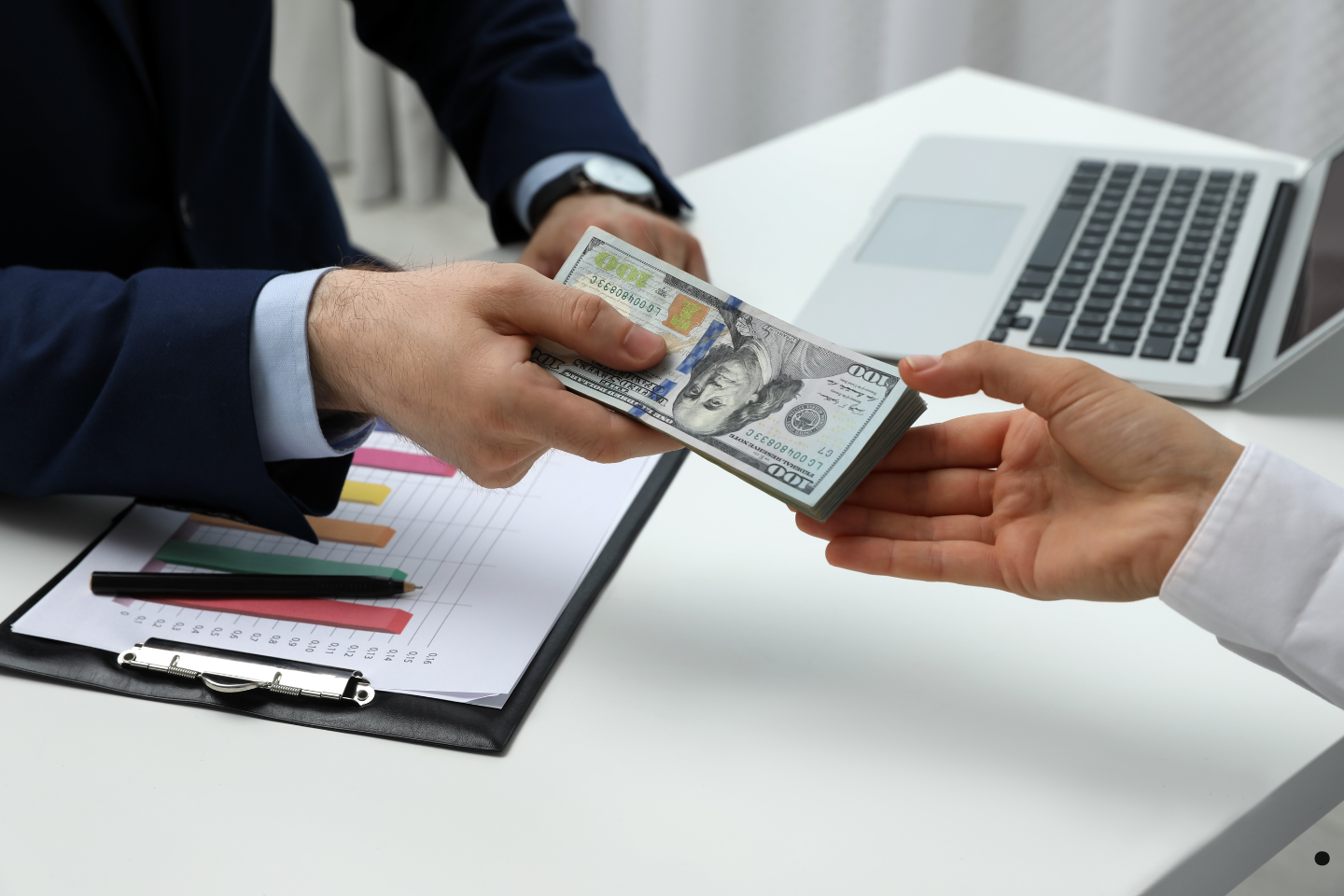 Businessperson handing over a stack of hundred-dollar bills across a desk with financial charts.