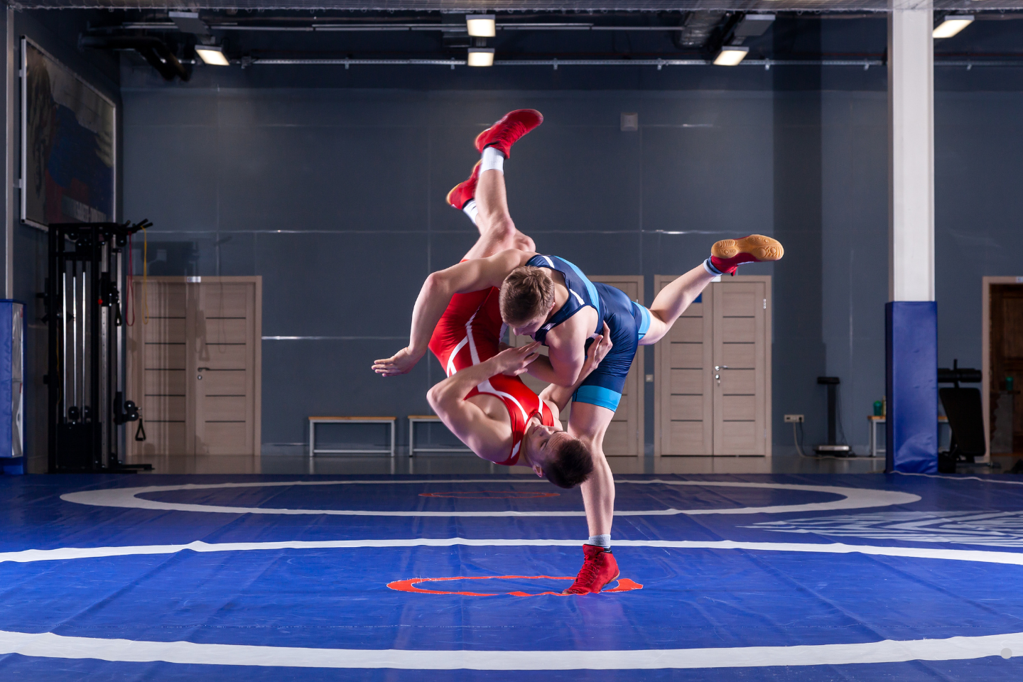 Wrestler in a blue singlet lifting and throwing an opponent in a red singlet inside a training facility.