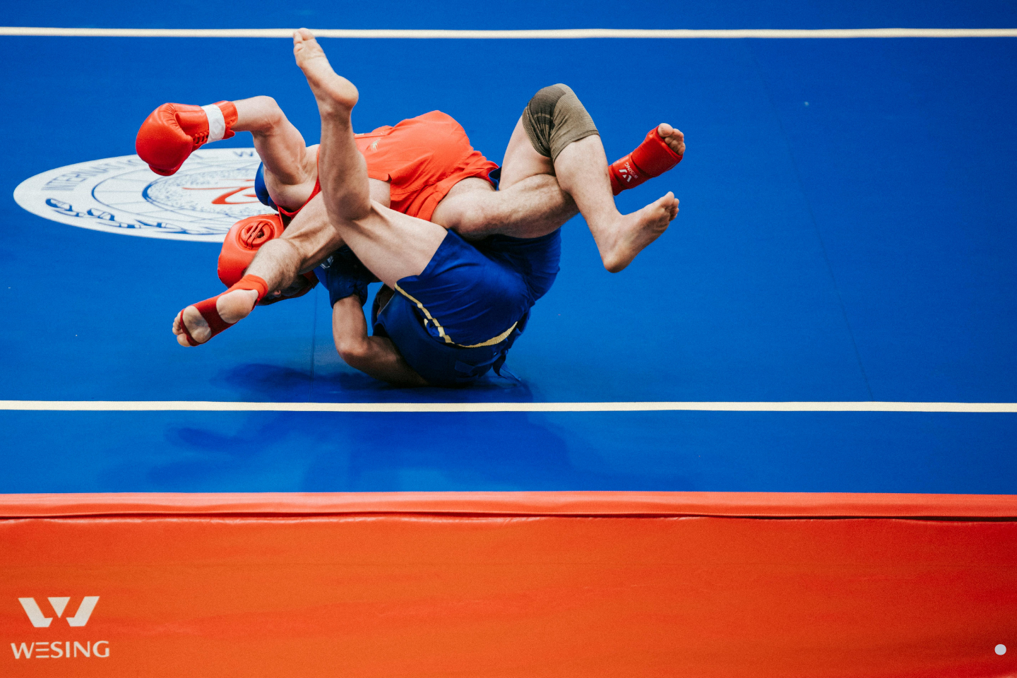 Two athletes grappling intensely on a blue competition mat during a wrestling match.