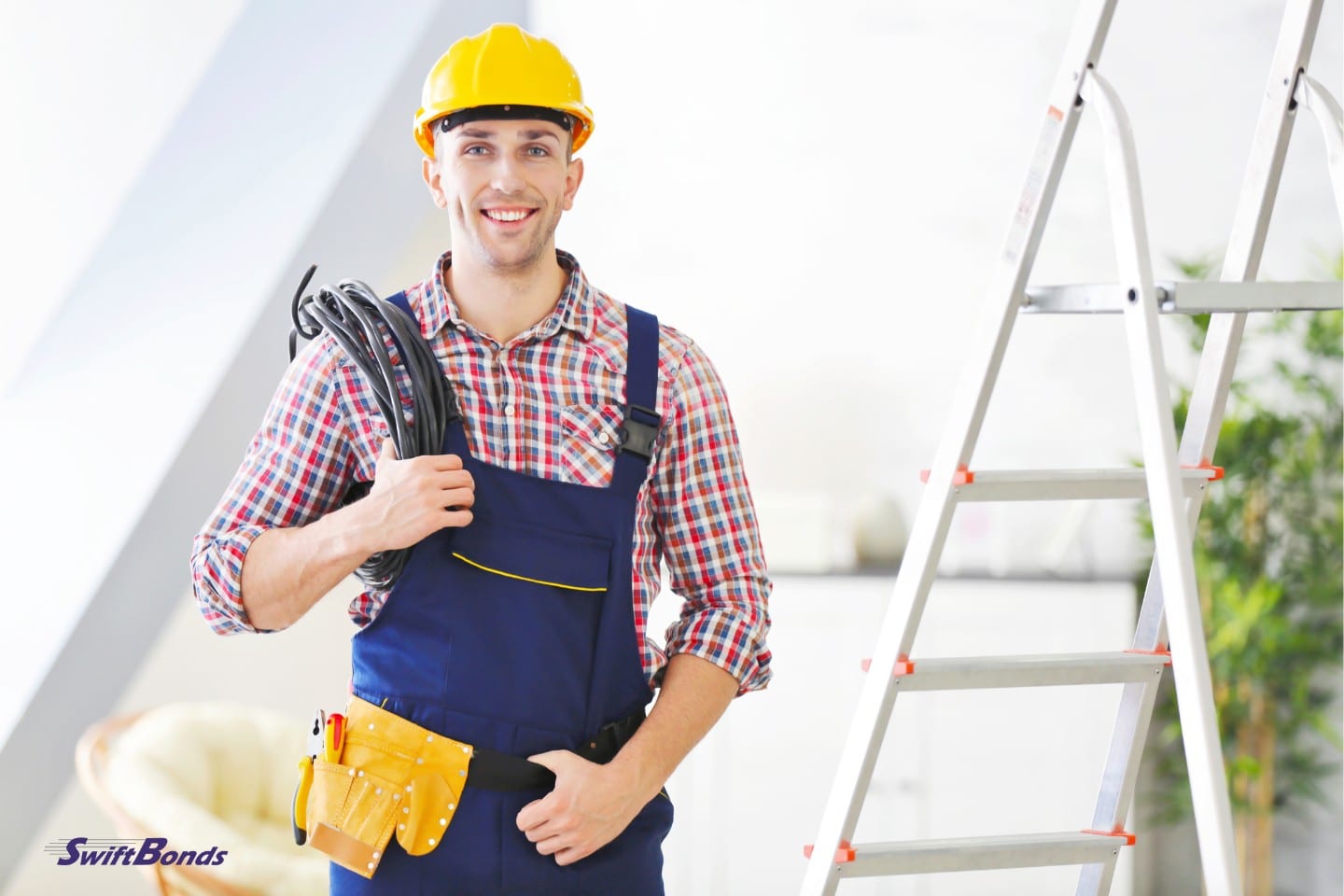An electrician wearing a yellow hard hat and holding tools stands near a stepladder.
