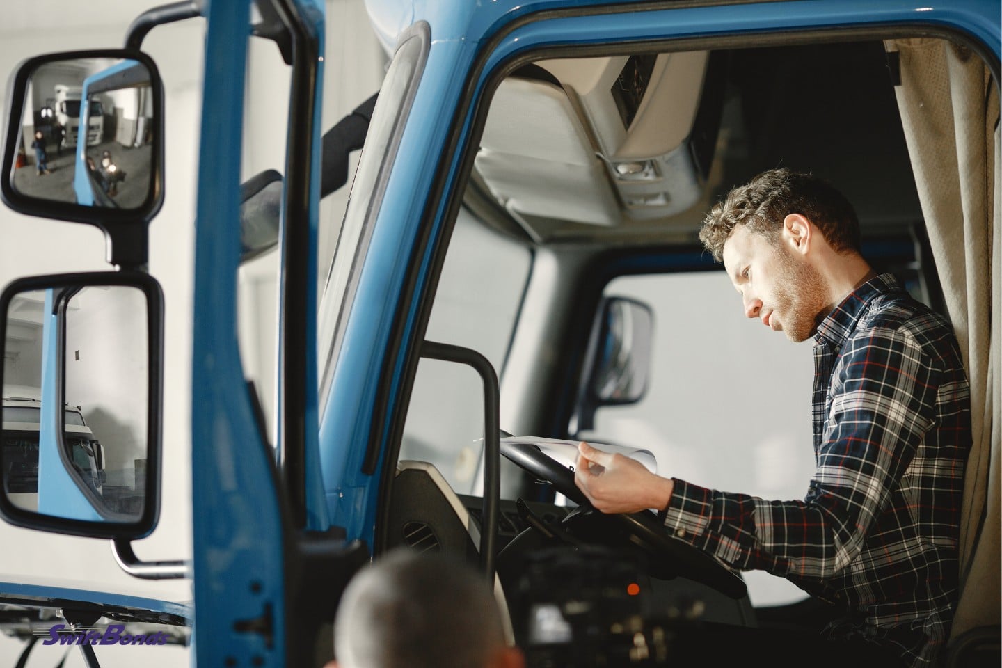 A man reading documents while seated in the body of a blue truck is wearing a plaid shirt.