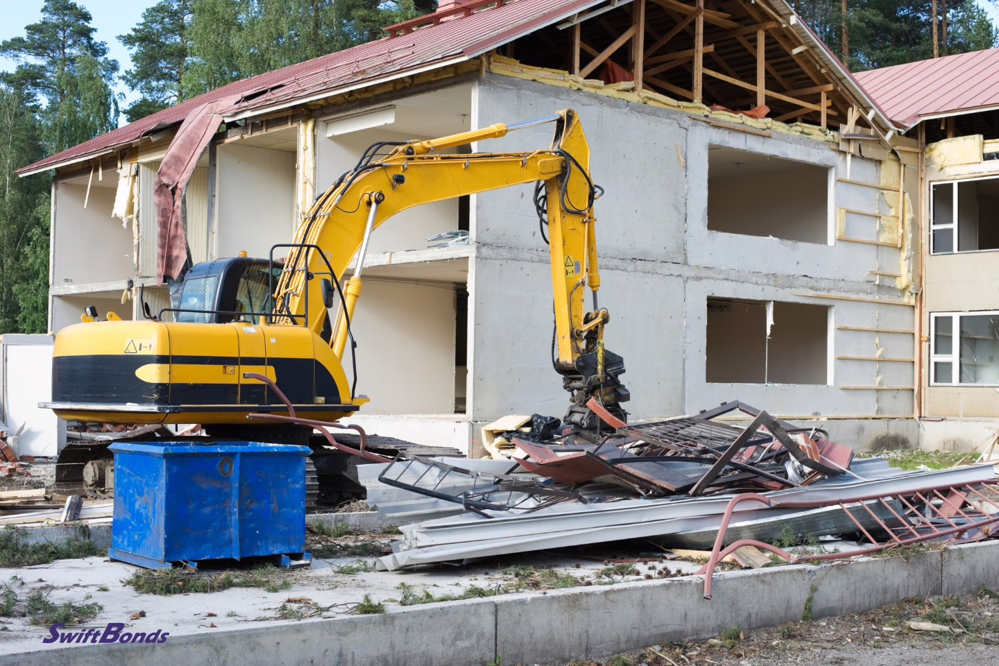 Scrap metal and an excavator stand in front of a half destroyed house.
